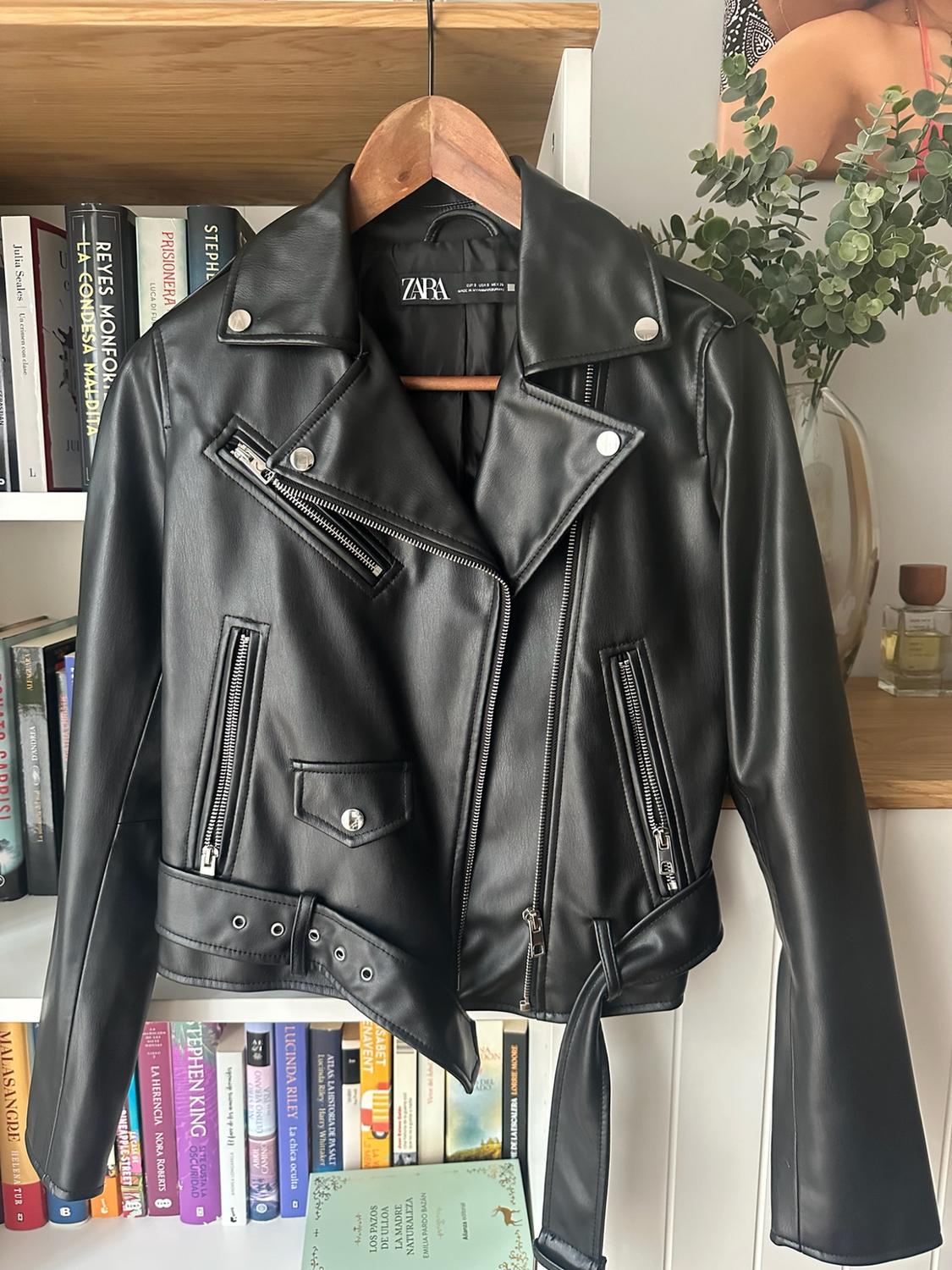 Black jacket with zips and a belt hanging on a hanger in front of a bookshelf. ZARA label.