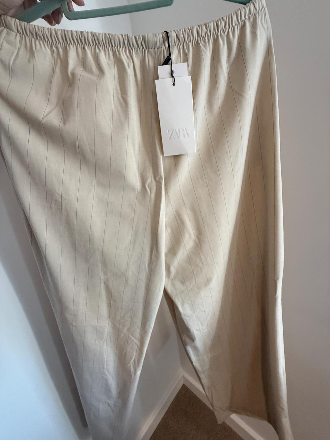 Hand holding a hanger with fine-striped beige trousers and an elasticated waistband. Label with Zara text. White wall background.