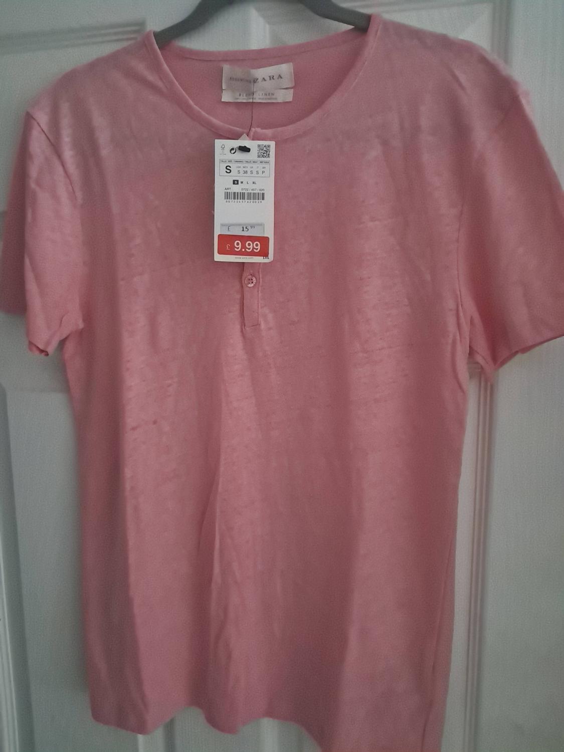 Pink short-sleeved button-up t-shirt, hanging on a white door. Label text: ZARA, S, 15.99, £ 9.99.