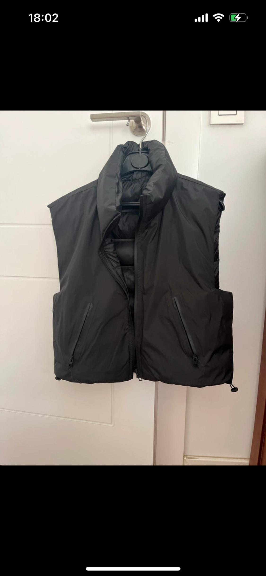 Black padded gilet hanging on a coat hook over a white door. The mobile phone screen displays the time 18:02.