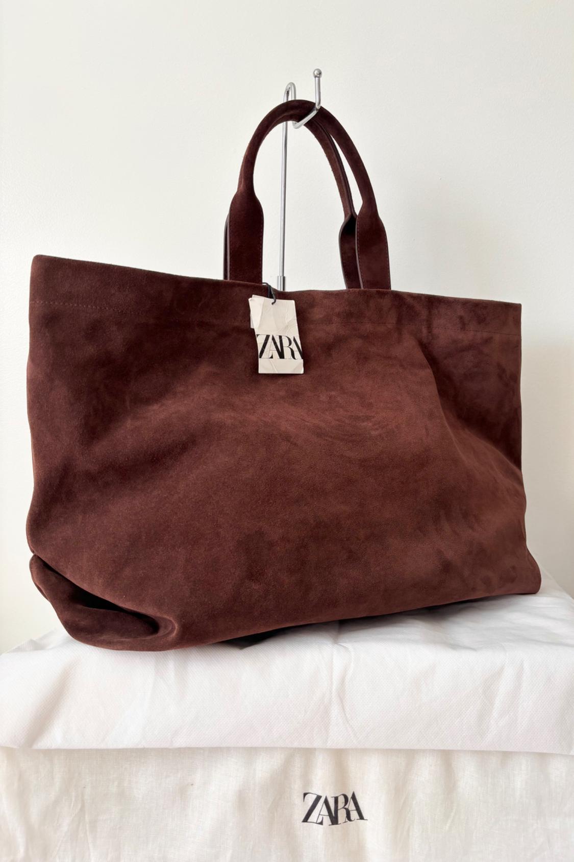 Large brown suede handbag with handles, hanging on a metal stand. Zara label visible. Minimalist white background.