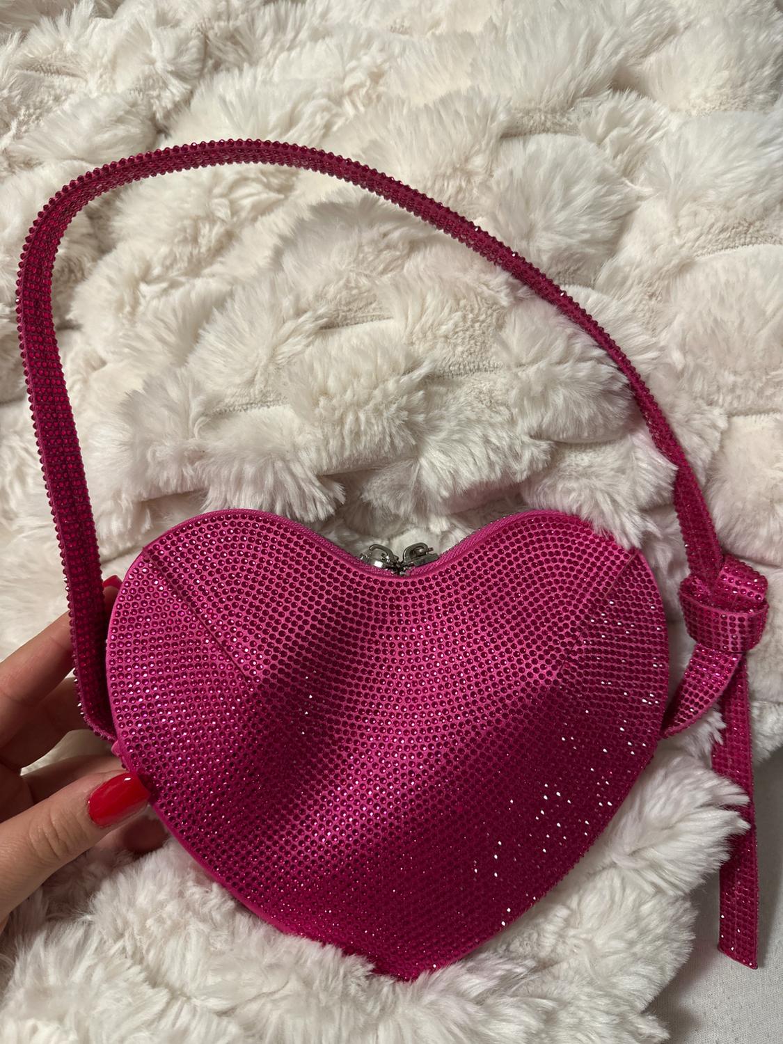 Shiny pink heart-shaped handbag with a handle, on a fluffy white fabric background. Hand with red nails holds the bag.
