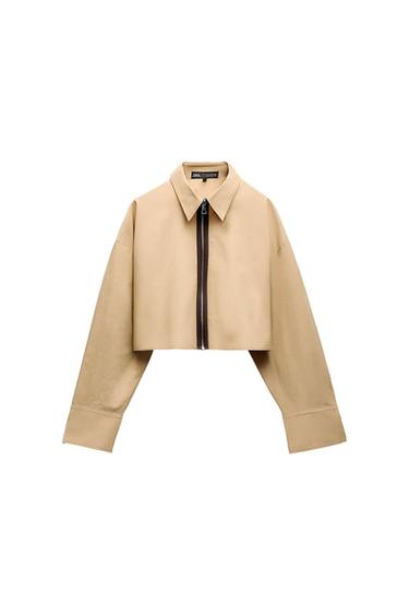 Beige cropped jacket with black front zip and classic collar. Visible label on the inside of the collar. White background.