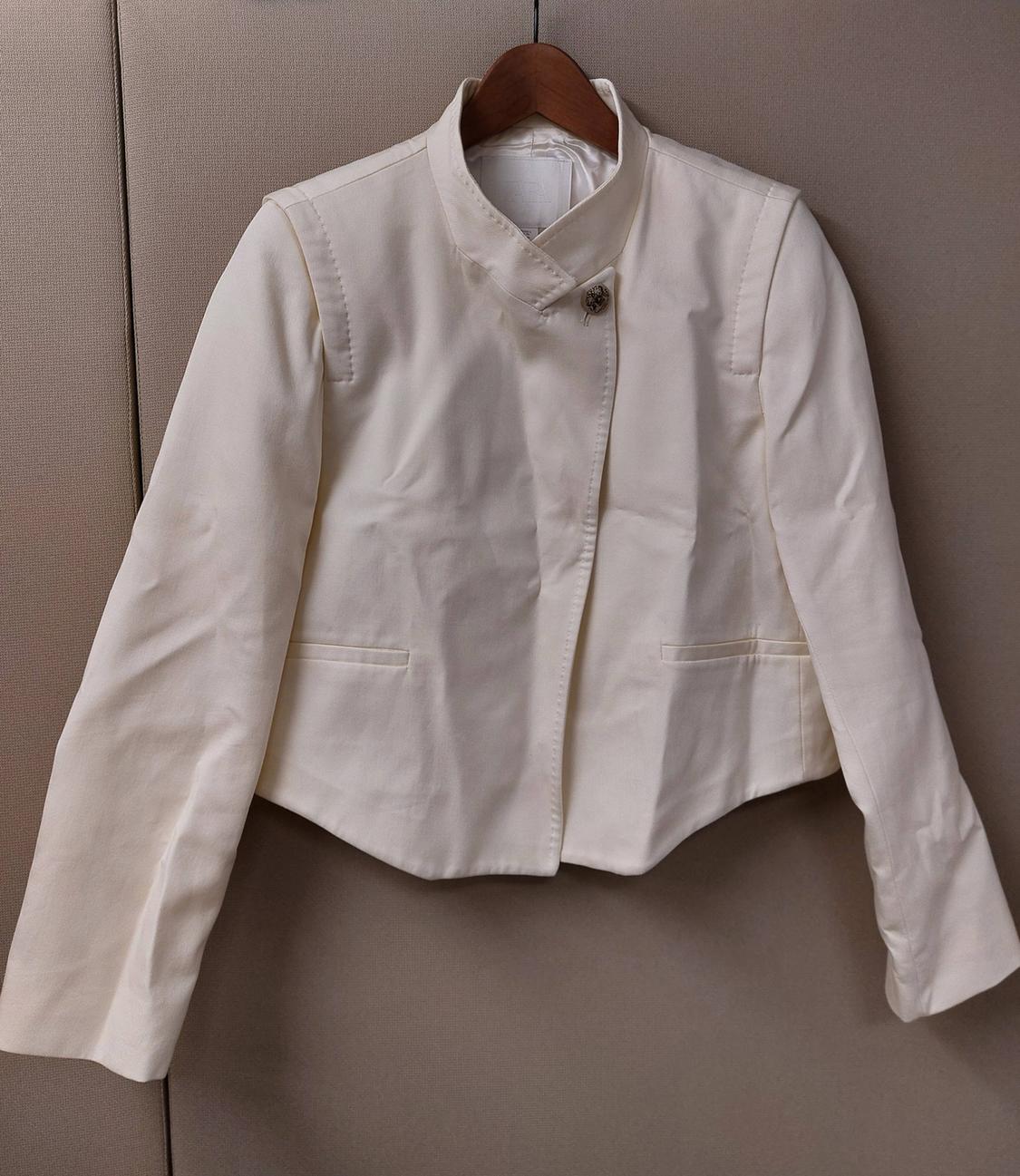 White cropped jacket hanging on a wooden hanger against a neutral background. Design featuring a high neck, asymmetrical one-button fastening, and long sleeves.