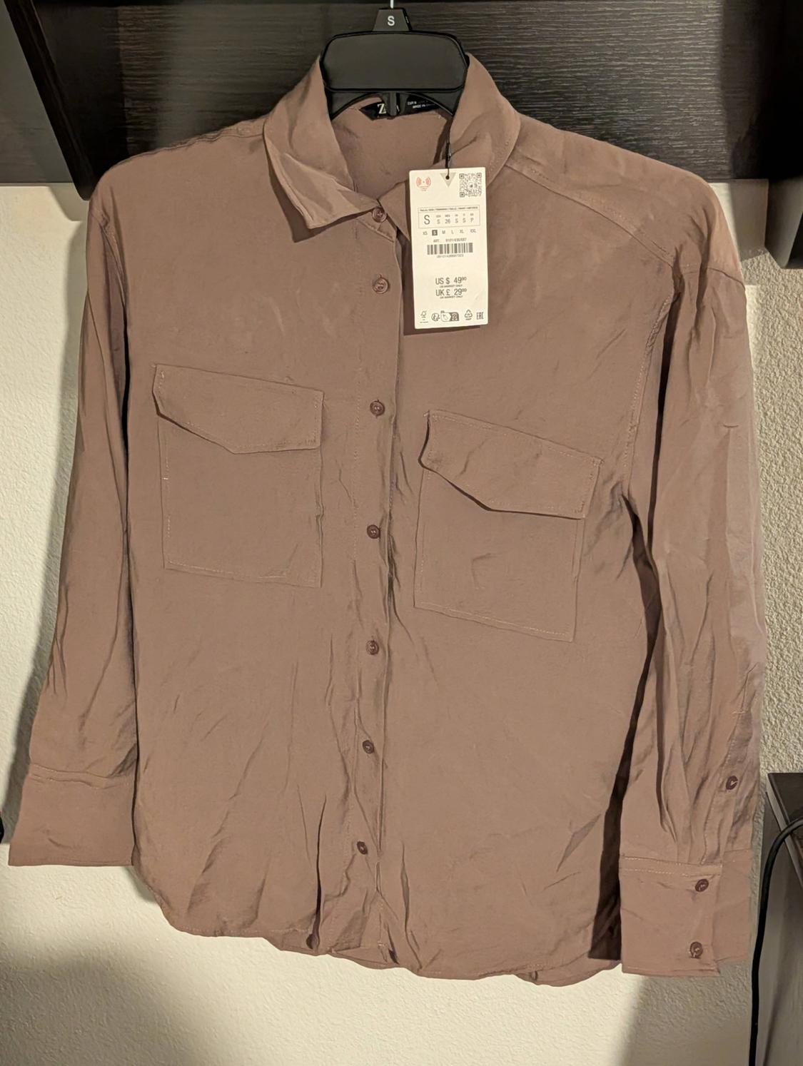Wrinkled brown shirt with front pockets, hanging on a black hanger. Visible tag with price and size S.