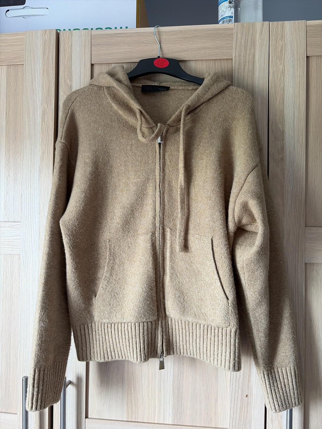 Beige ZARA knitted hooded sweatshirt with zip, hanging on a hanger in front of a light wood wardrobe. Casual and relaxed style.