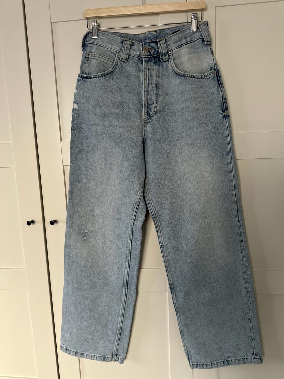 Ανοιχτόχρωμο ψηλόμεσο τζιν κρεμασμένο με μανταλάκια σε ξύλινη κρεμάστρα μπροστά από τις λευκές πόρτες μιας ντουλάπας.