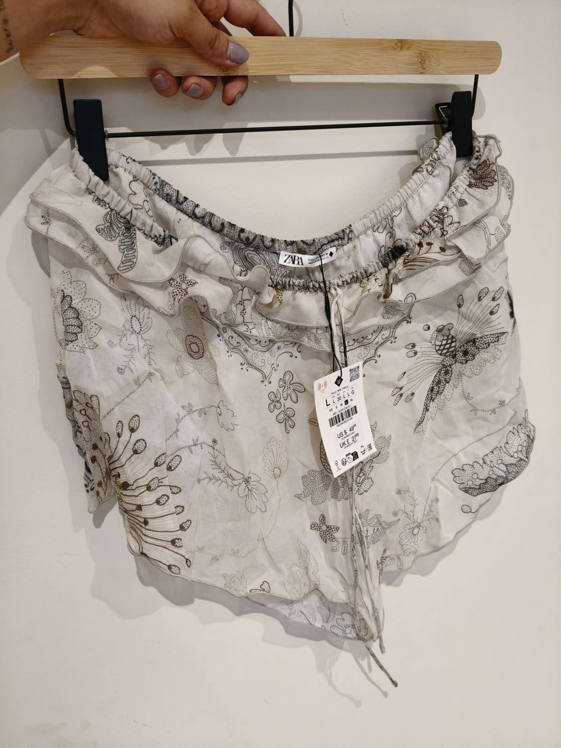 Hand holding a hanger with white ZARA shorts. They feature a natural print, an elasticated waist and ruffled hems.