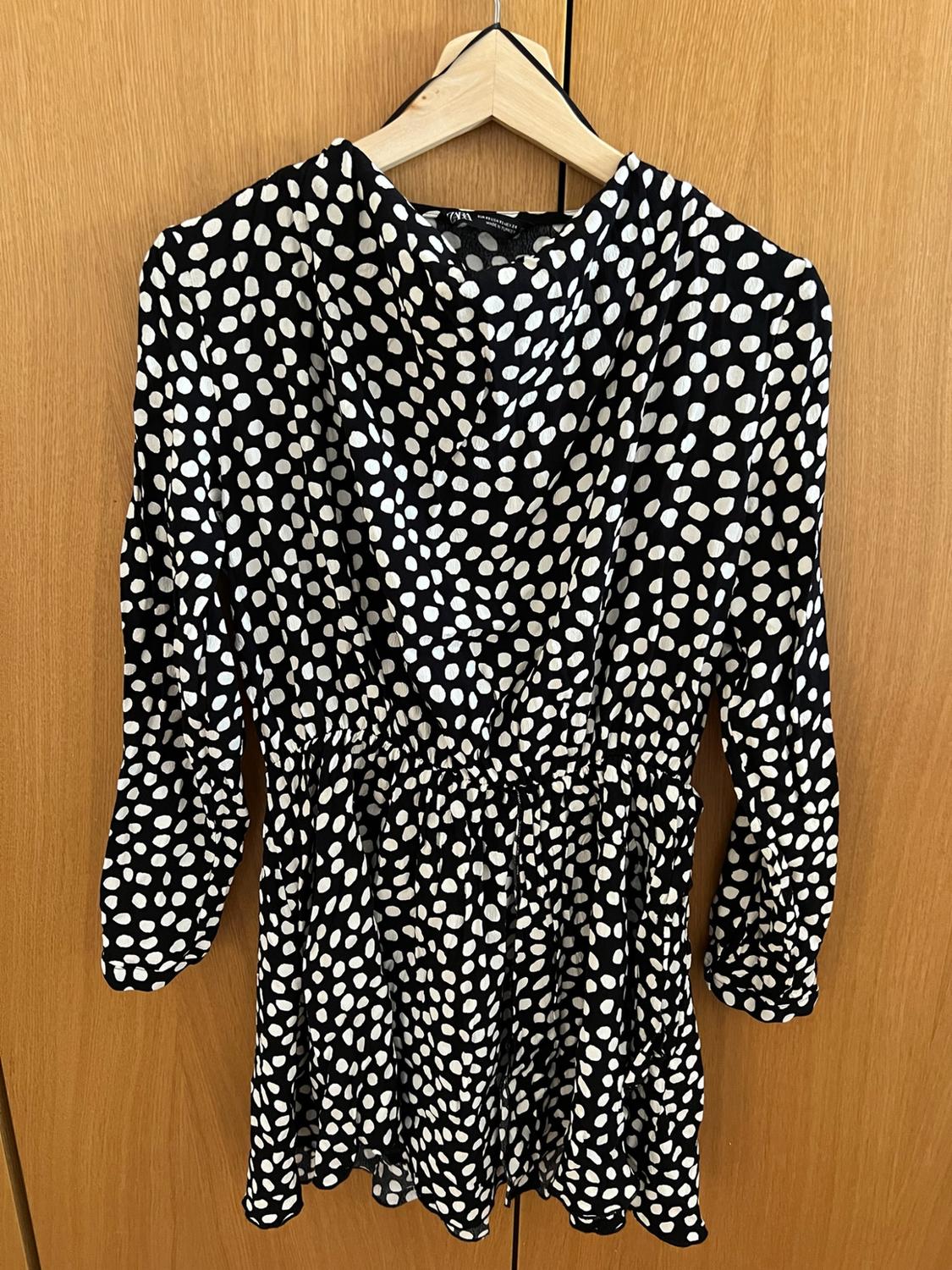 Black polka-dot long-sleeved dress hanging on a hanger against a wooden background. Visible text: ZARA.
