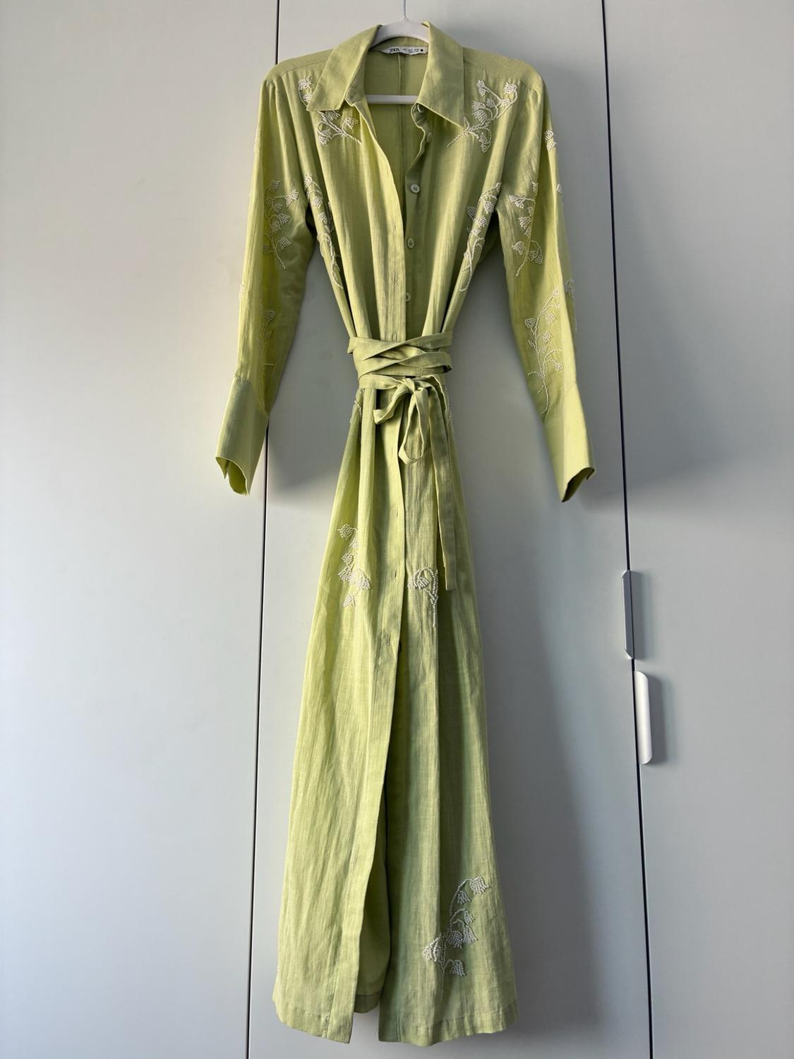 Long, light green shirt dress with white floral embroidery and a tie belt, hanging on a hanger in a white wardrobe.