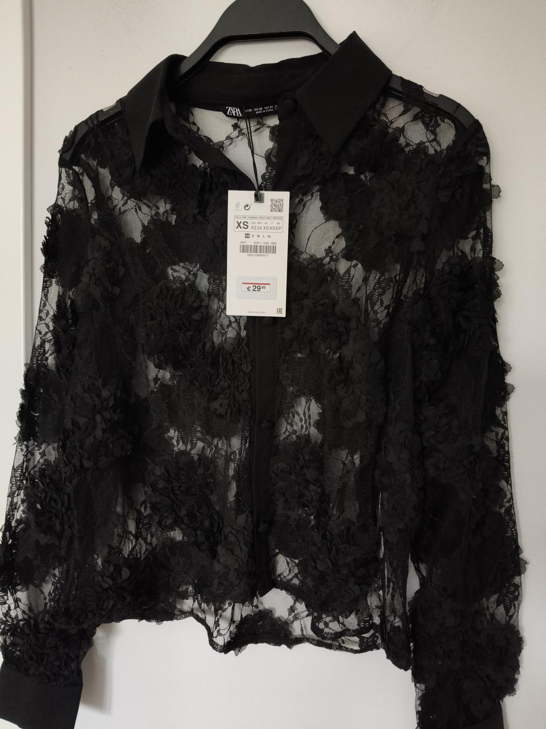 Black floral lace blouse hanging on a hanger, with a visible price tag of €29.95 and size XS.