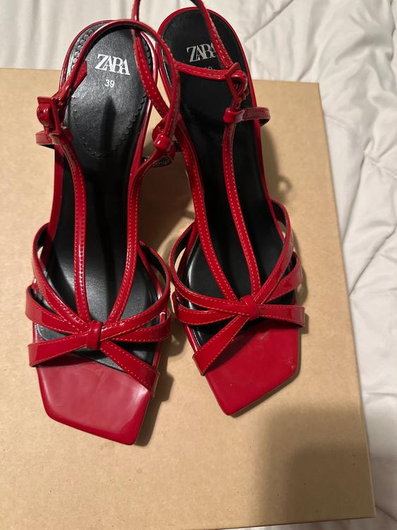 Red high-heeled sandals with criss-cross straps and buckles, on a brown box. Visible text: "ZARA 39".