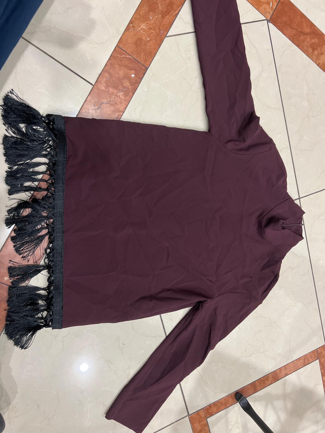 Burgundy long-sleeved dress with black fringe on the hem, placed on a beige and brown tiled floor.