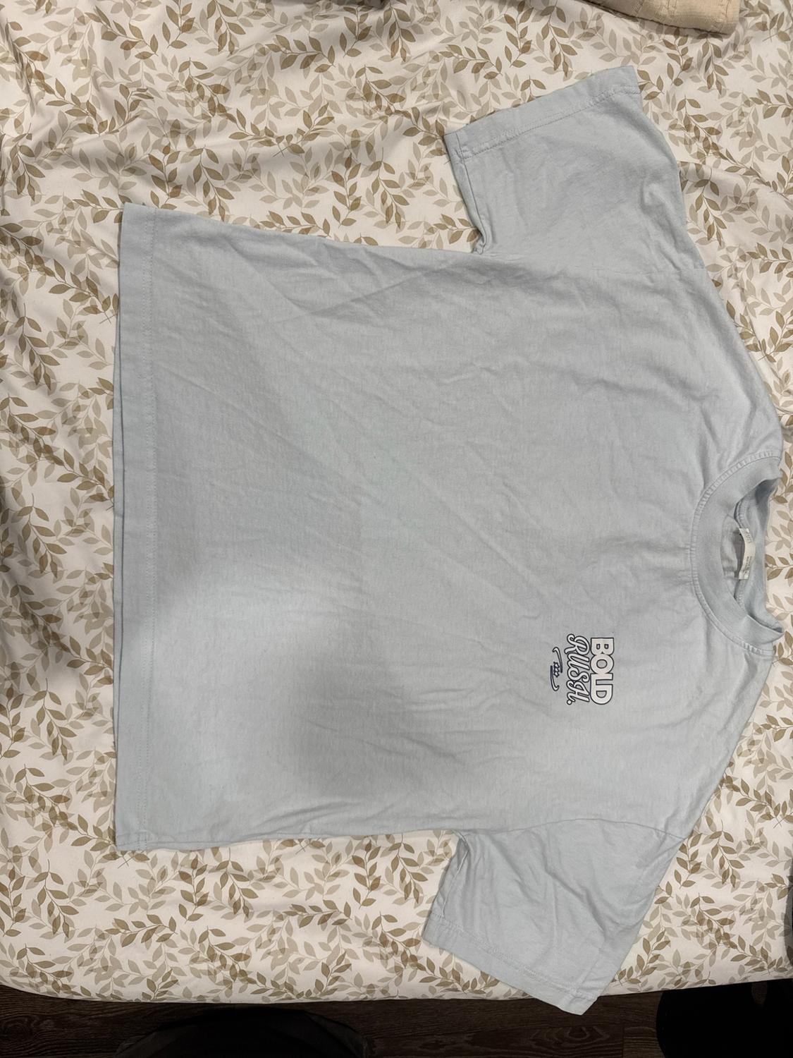 Light blue short-sleeved t-shirt against a leaf-print background. BOLD RUSH text detail on the chest. Casual style.
