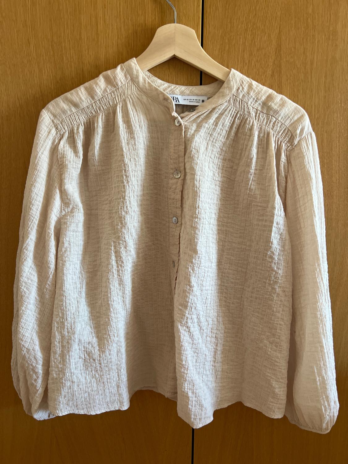 Beige, relaxed-fit long-sleeved blouse with buttons and textured fabric, hanging on a wooden hanger in front of a wardrobe.