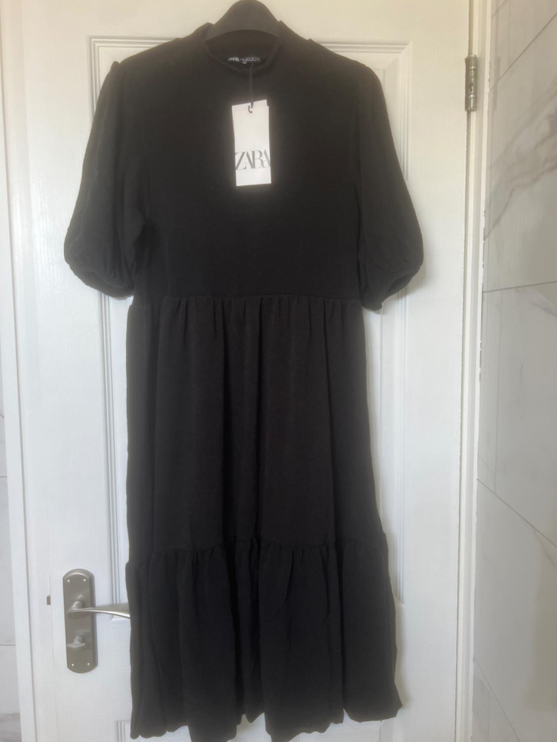 Short-sleeved black dress hanging on a white door. Visible label with the text "ZARA". Illuminated interior setting.
