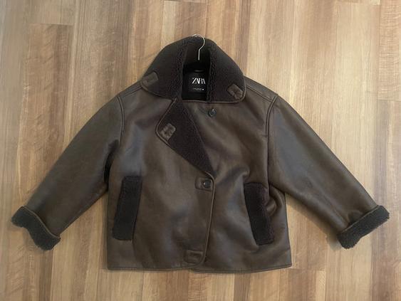 Brown jacket with dark lining and a wide collar, hanging on a hanger over a wooden floor. Visible tag: ZARA.