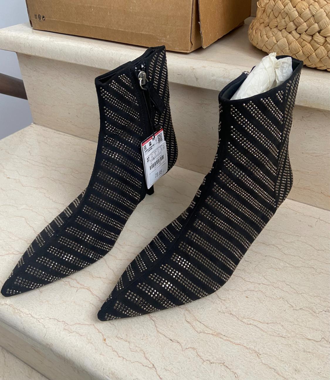 Front view of black ankle boots with a perforated stripe design and side zip, with a visible price tag.