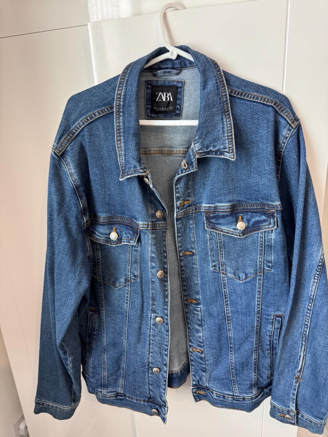 Blue denim jacket hanging on a white hanger. Label visible on the collar. Plain white background.