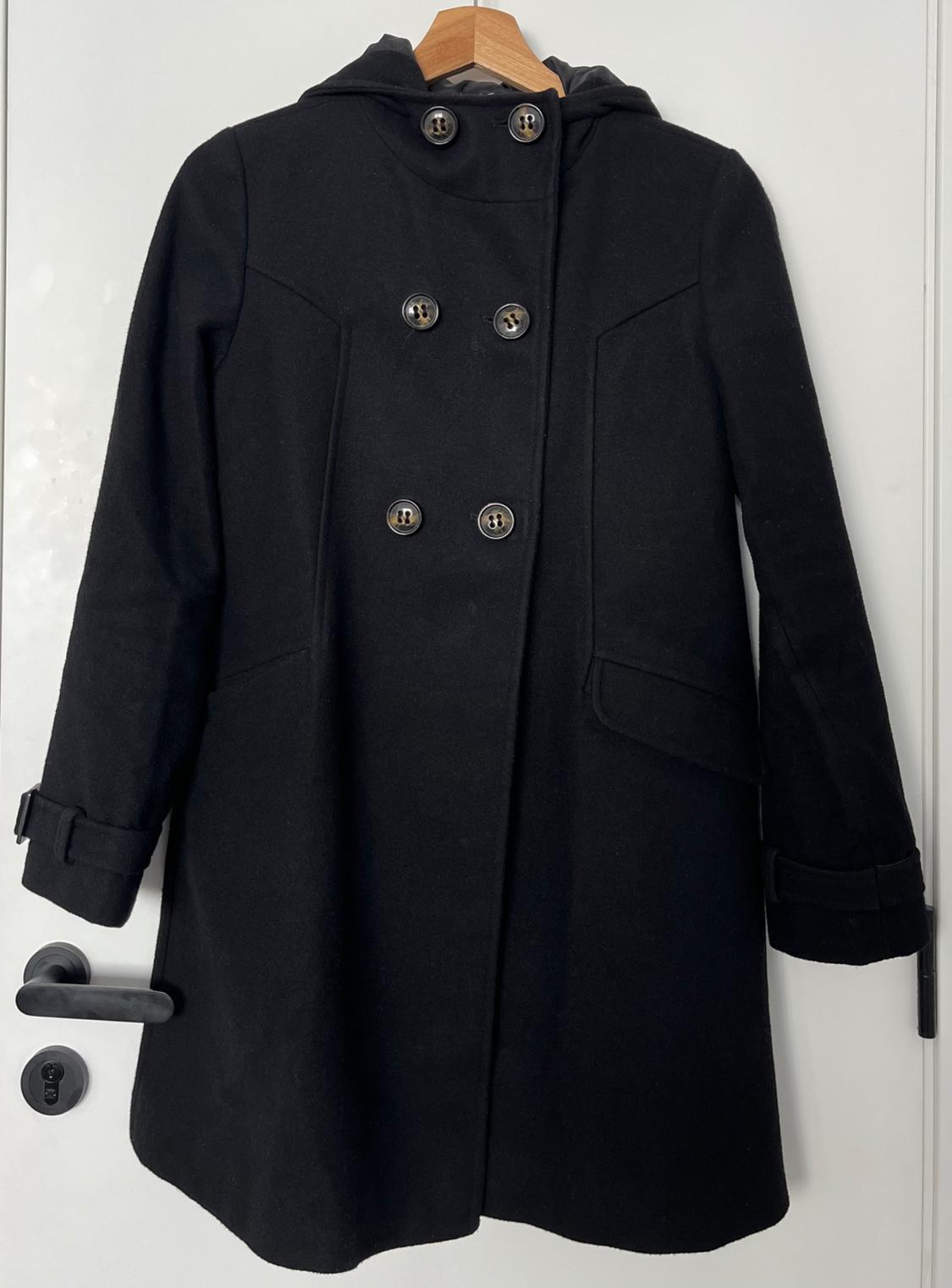 Black wool coat with a hood and double-breasted fastening, hanging on a wooden hanger over a white door.