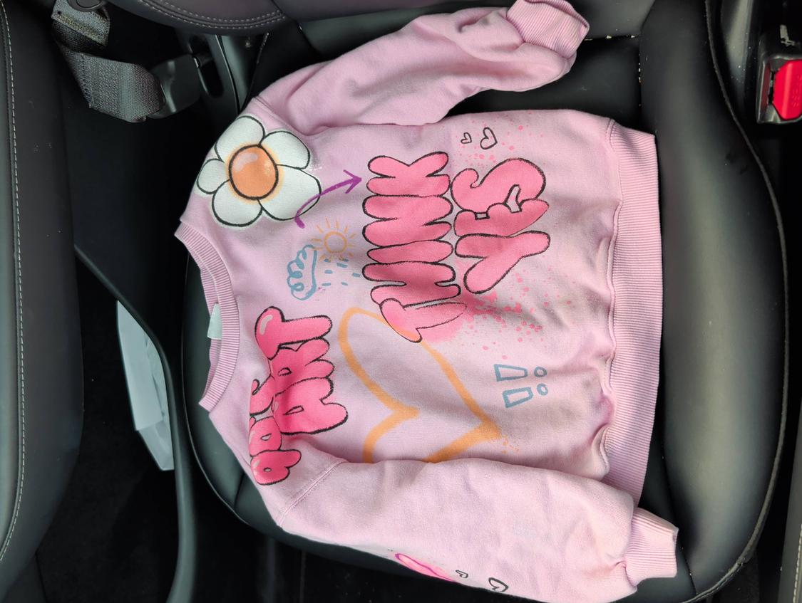 Pink sweatshirt with graphic design of a flower, heart, and the text "Stay Happy" in large letters, on a black car seat.