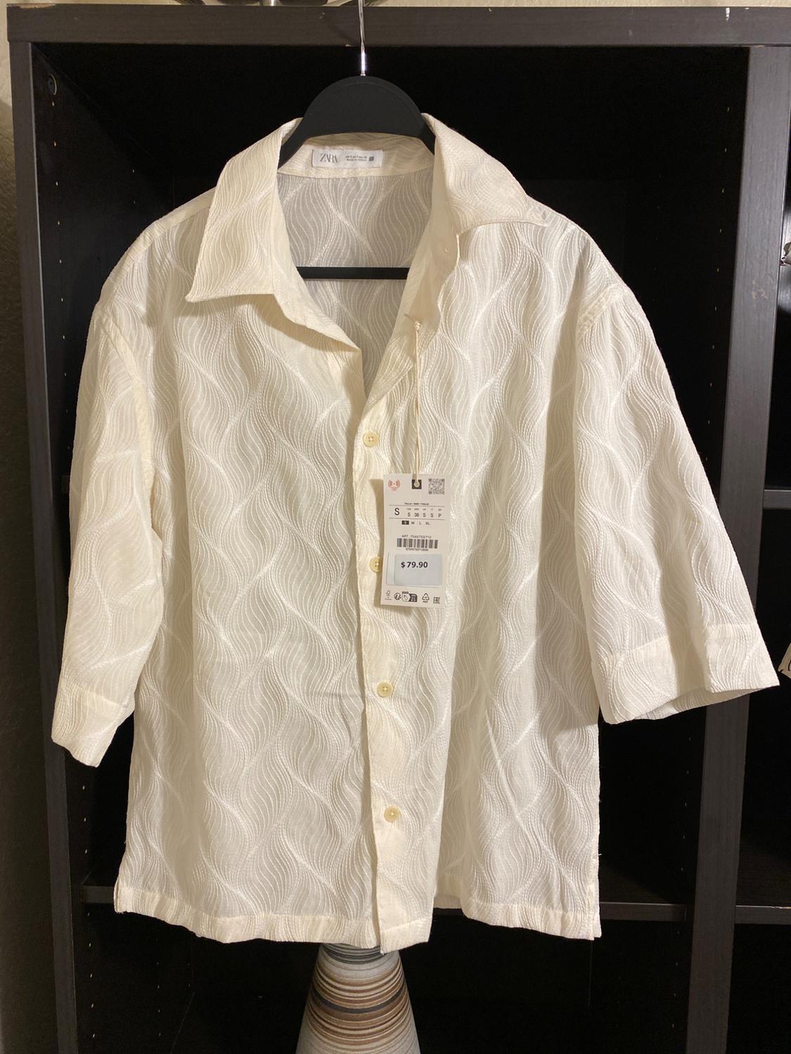 White shirt with a wavy texture hanging on a black hanger. Visible tag with a price of $79.90. Dark shelving in the background.
