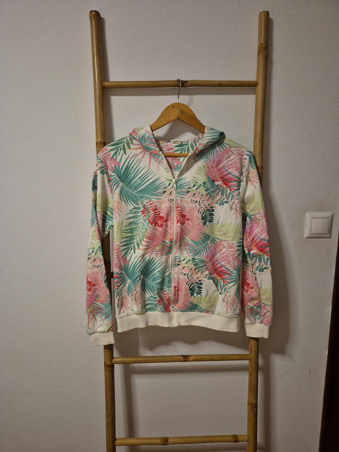 White zip-up sweatshirt with a tropical print of pink and green leaves, hanging on a bamboo ladder against a white wall.