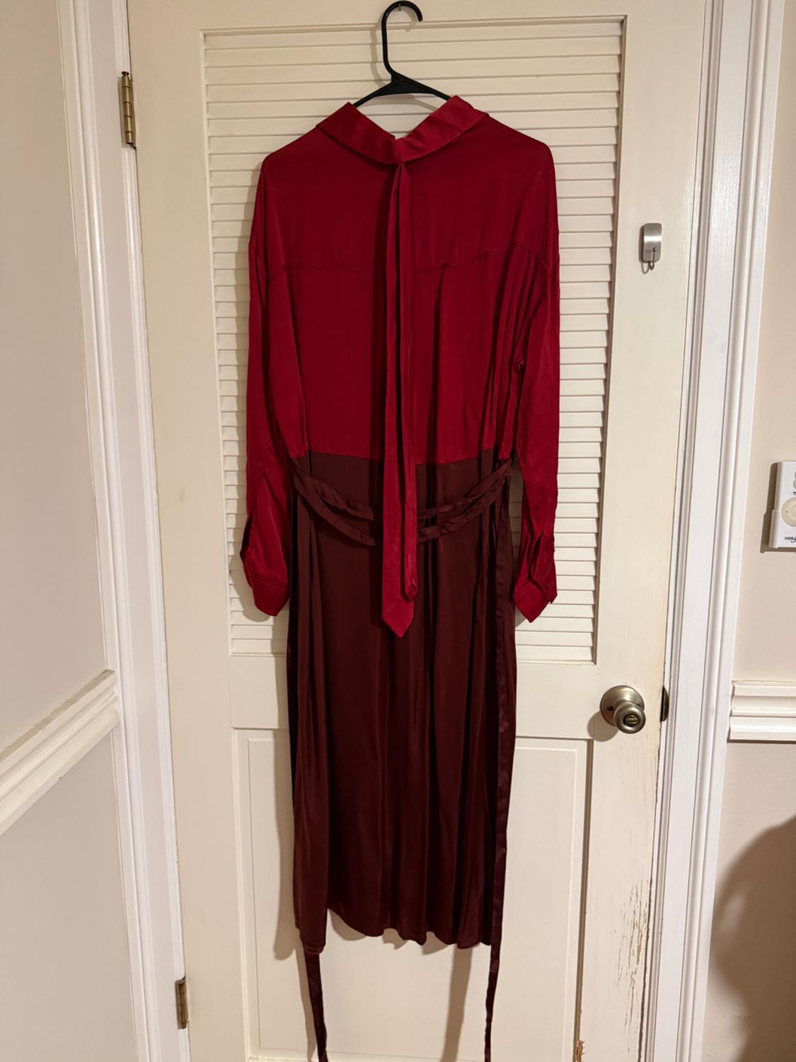 Red and brown dress hanging on a white door. It has a bow at the neck and long sleeves. Indoor setting.