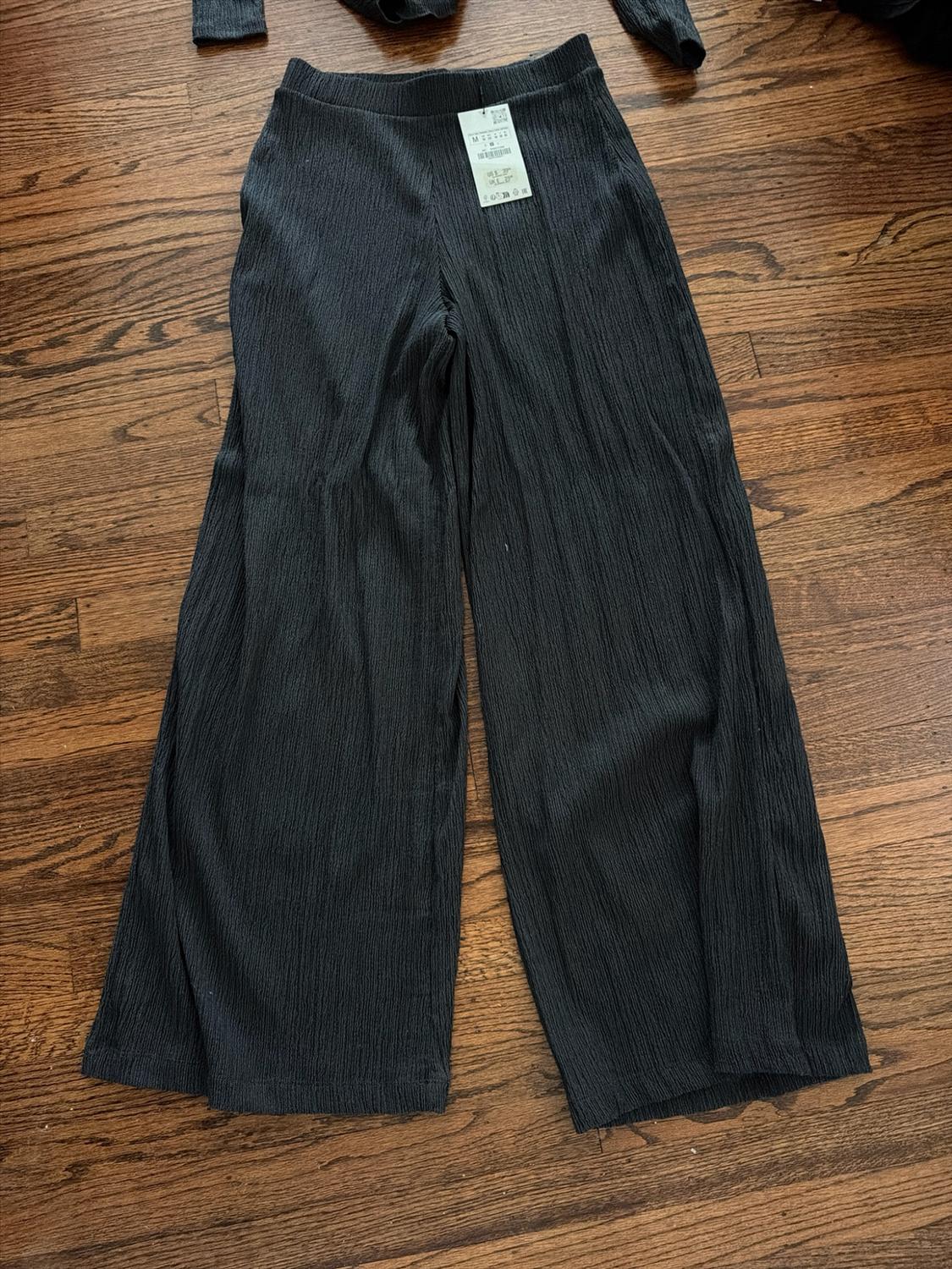 Black pants with wide legs and wrinkled texture, with a price tag, placed on a wooden floor.