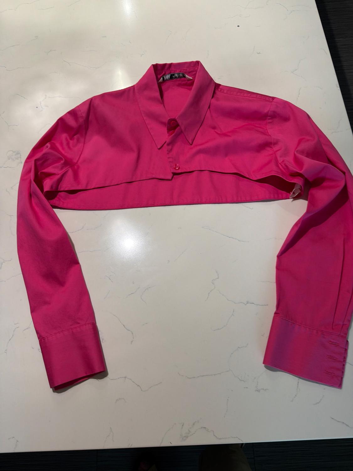 Cropped long-sleeve fuchsia pink shirt with a collar and buttons, laid out on a white marble surface.