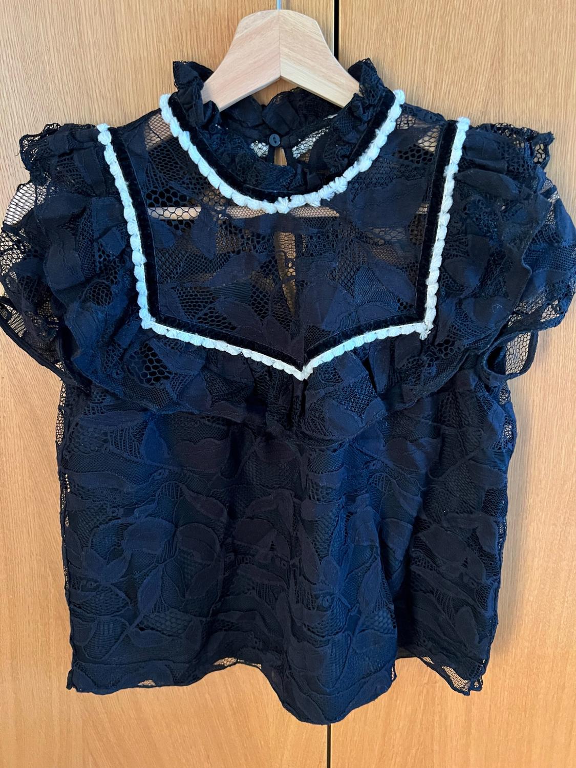 Black floral lace high-neck blouse with short sleeves and contrasting white trim. Hanging on a coat hanger against a wooden background.