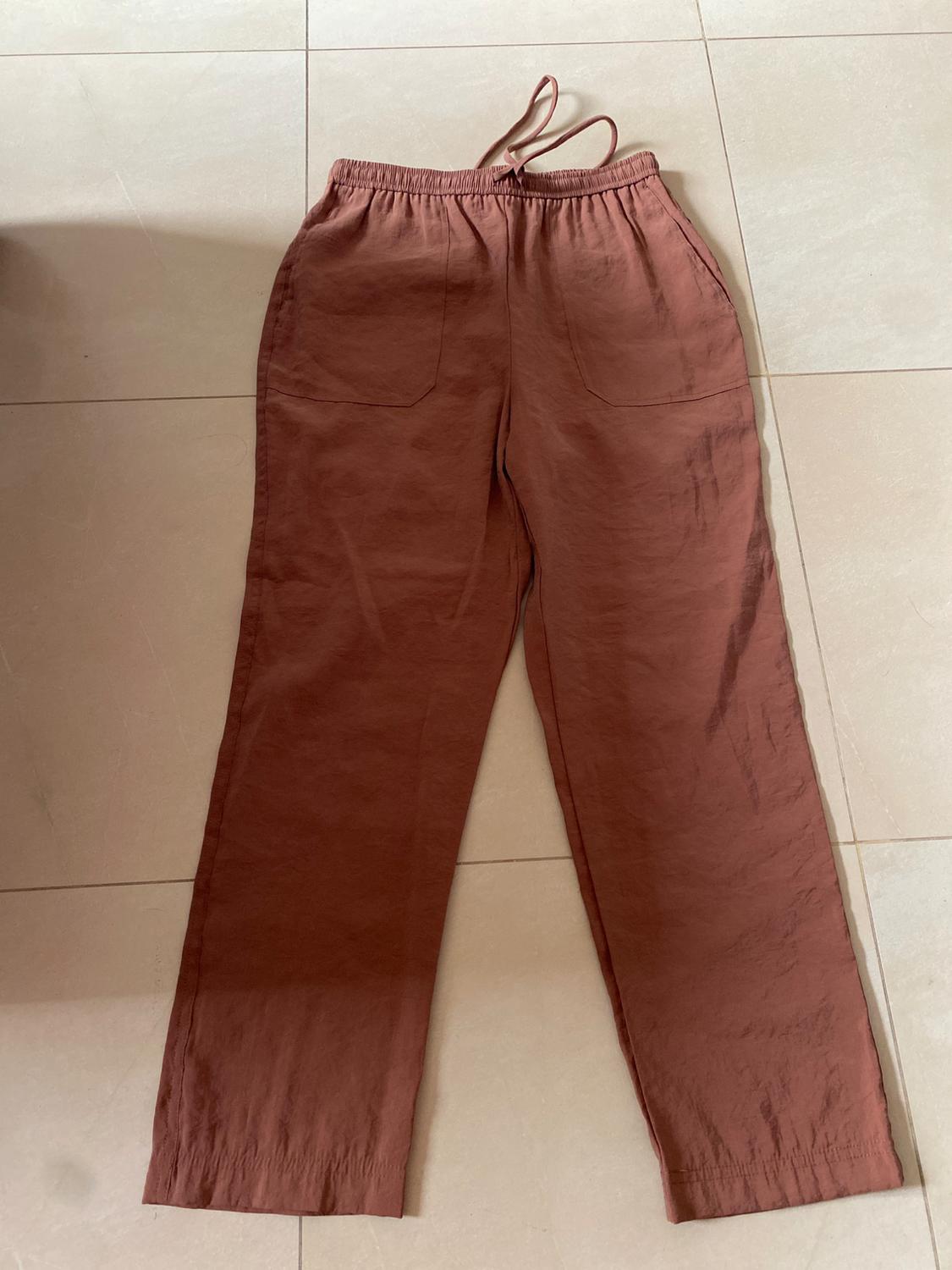 Brown long trousers with an elasticated waist and drawstring, placed on a light tiled floor.