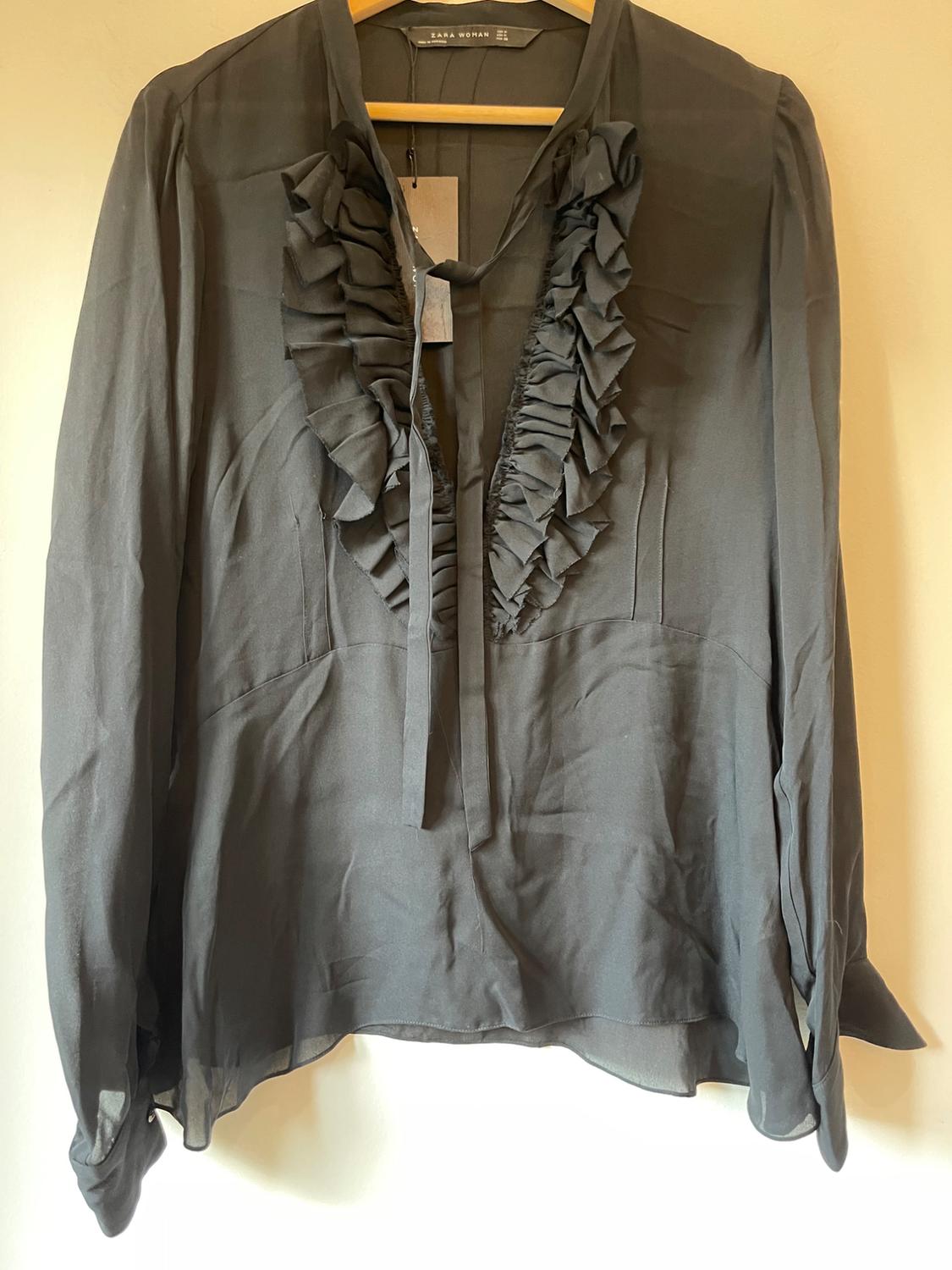 Black blouse with frills on the chest, hanging on a wooden hanger. Label visible on the neck that says "Zara Woman".
