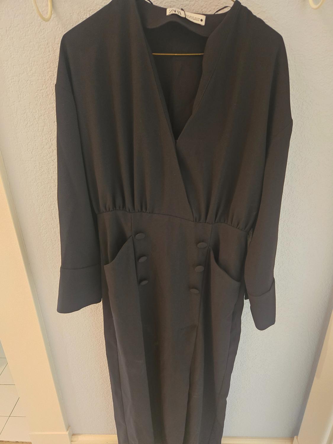 Black dress hanging on a hanger, with a V-neck and decorative buttons on the front. Light-coloured wall background.