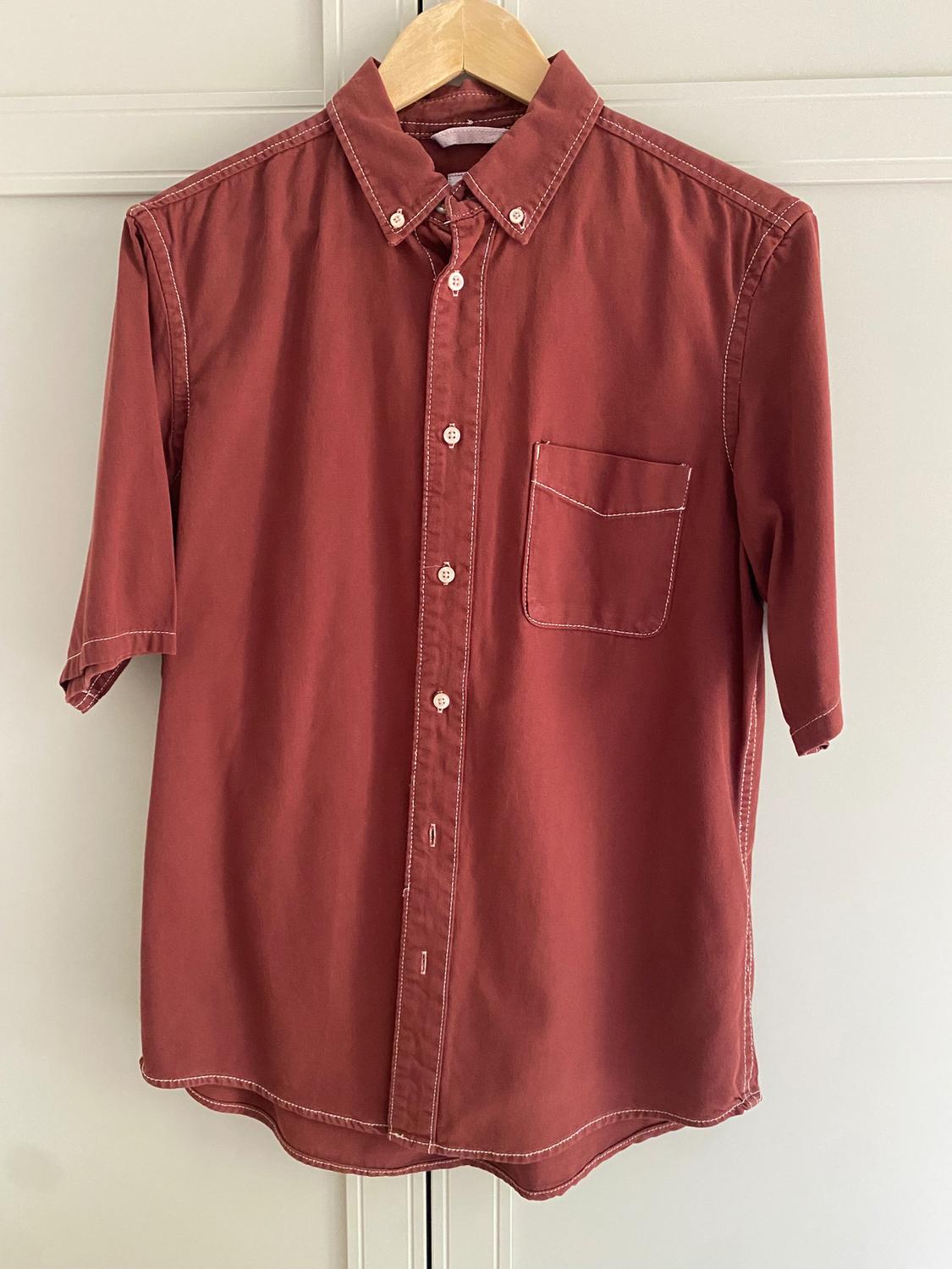 Dark red, short-sleeved shirt hanging on a wooden hanger, with a chest pocket and visible seams.