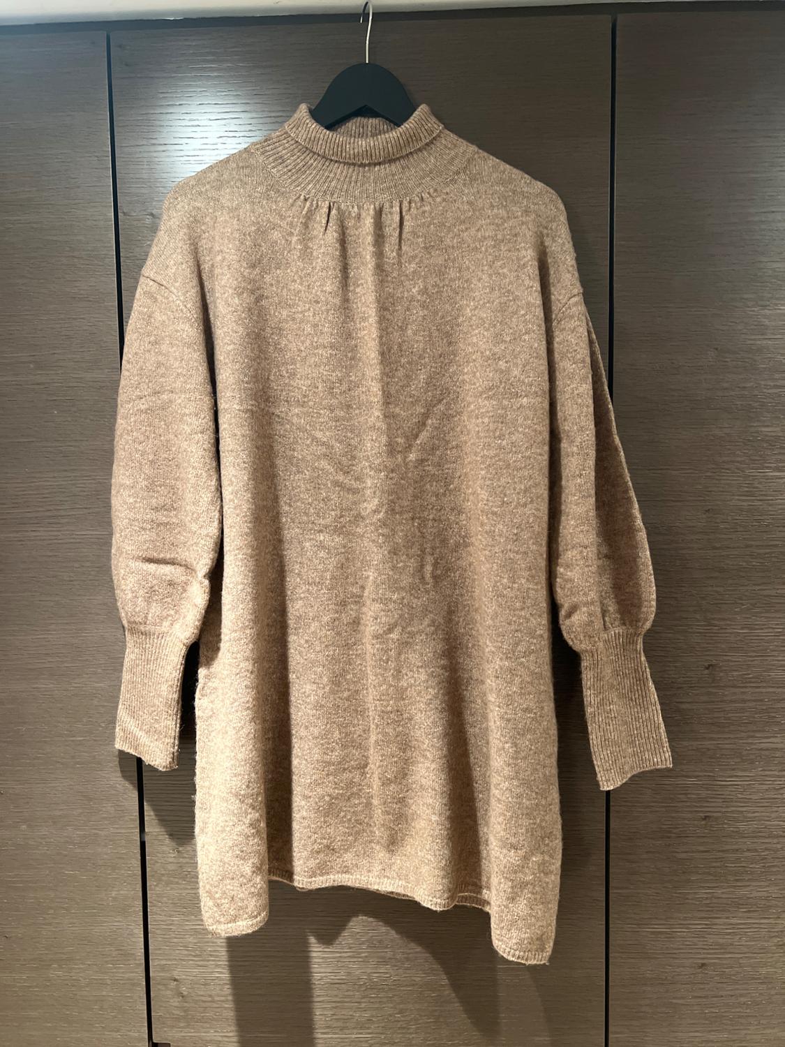Beige roll-neck jumper hanging on a black hanger, against a dark wooden background. Long sleeves and soft knit fabric.