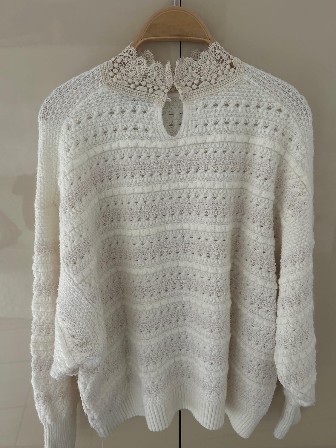 White knitted jumper with a lace collar, hanging on a wooden hanger, light beige background.