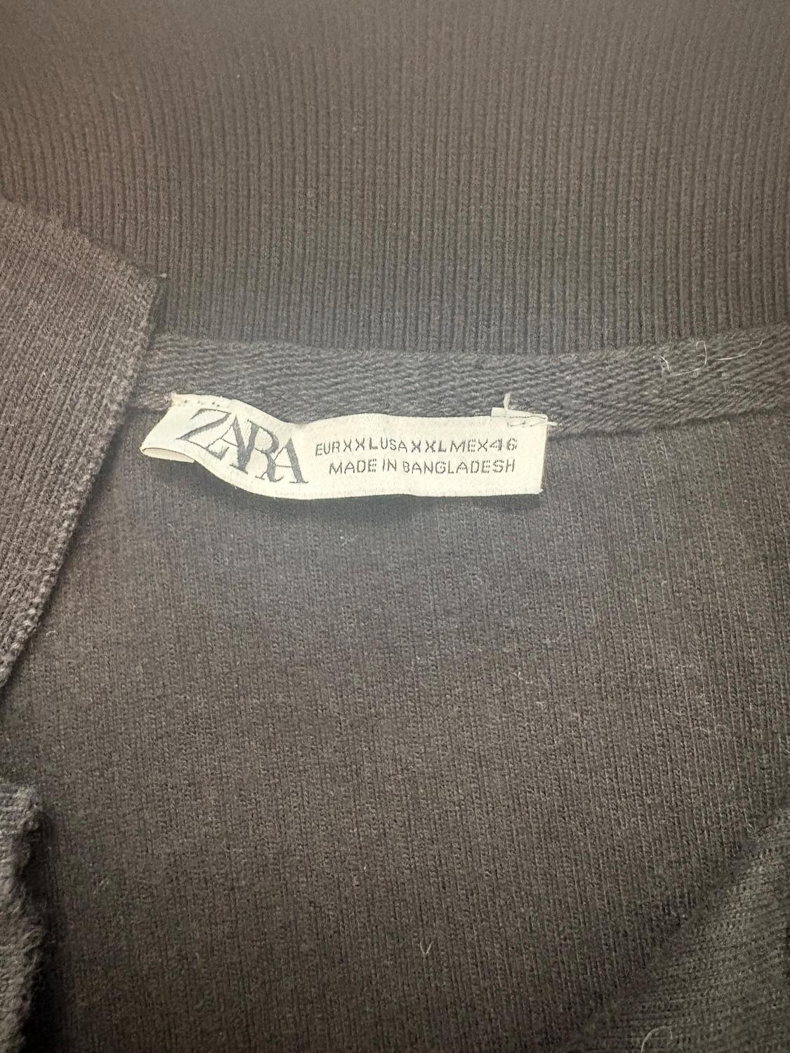 Black clothing label with text: "ZARA, EUR XXL USA XXL MEX 46, MADE IN BANGLADESH". Knit fabric visible around the label.