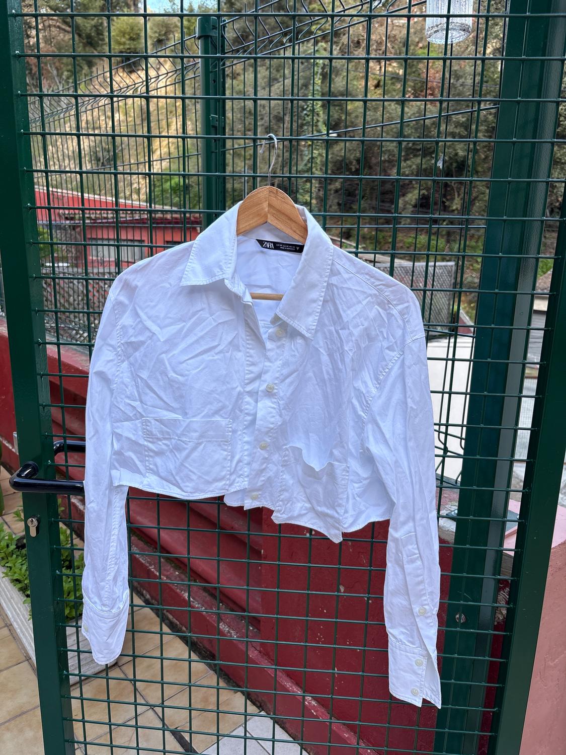 White short shirt hanging on a wooden hanger over a green fence, with a garden background and red wall.