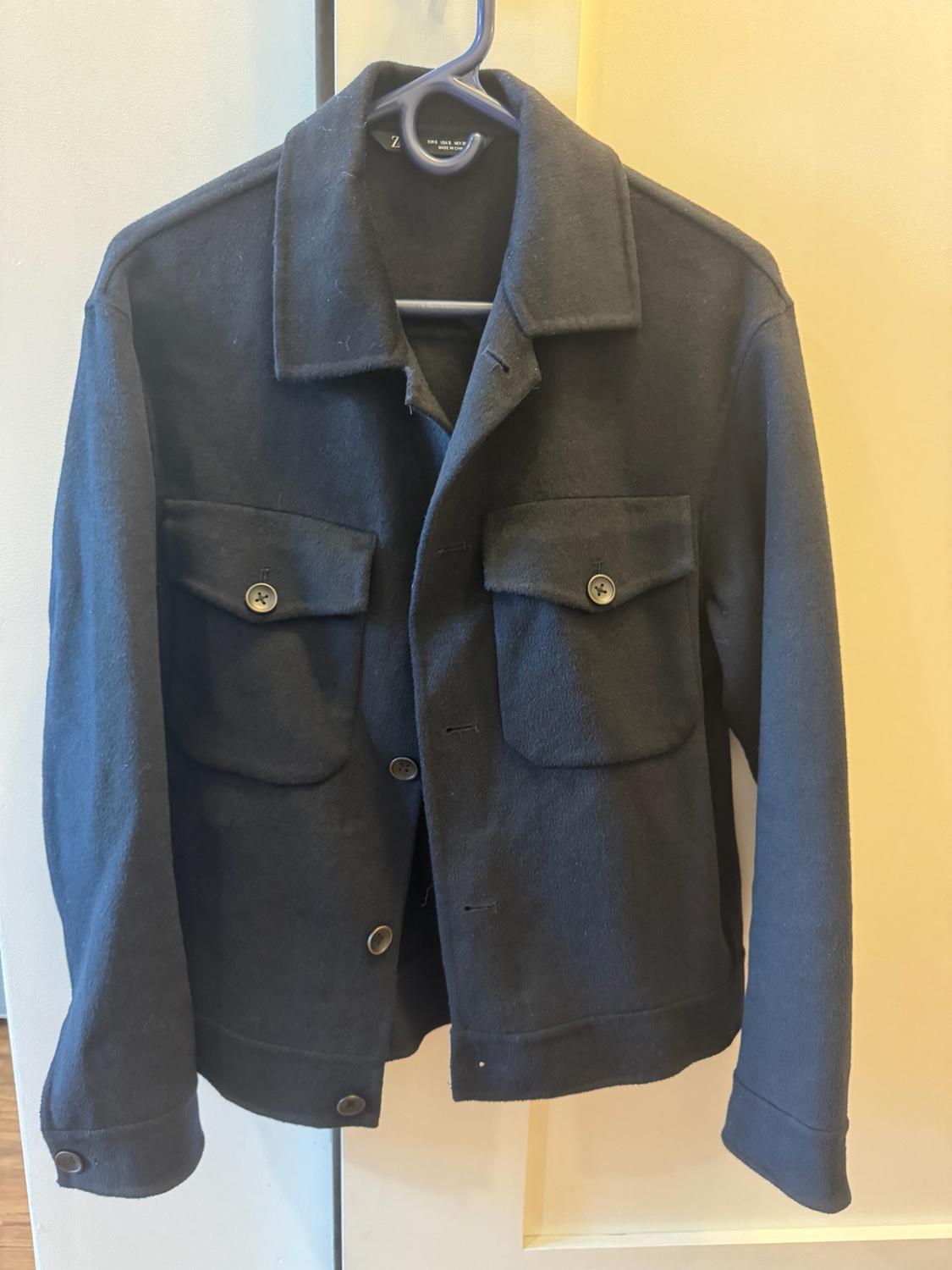 Black long-sleeved jacket with front buttons and two flap pockets on the chest, hanging on a blue hanger against a light background.