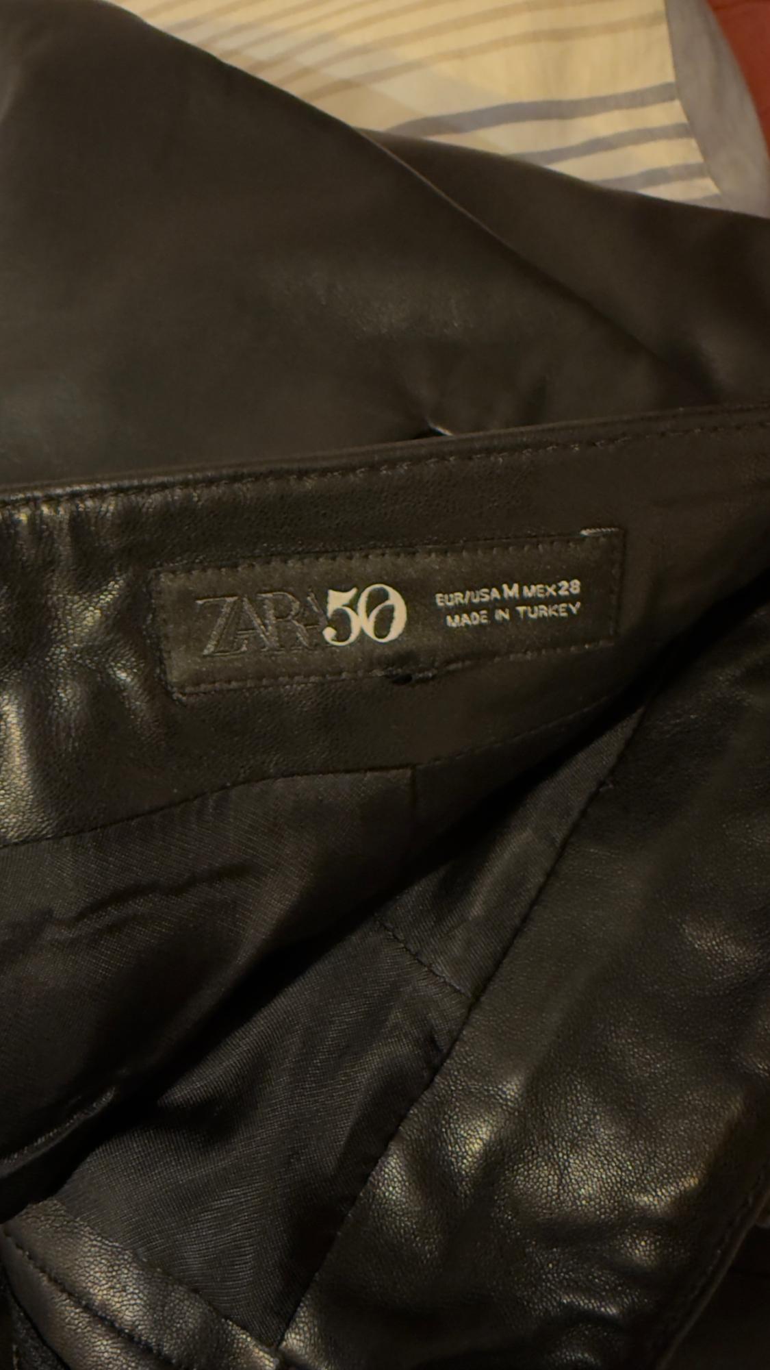 Black jacket label with text: "ZARA 50 EUR/USA M MEX 28 MADE IN TURKEY". Background fabric with grey and beige stripes.