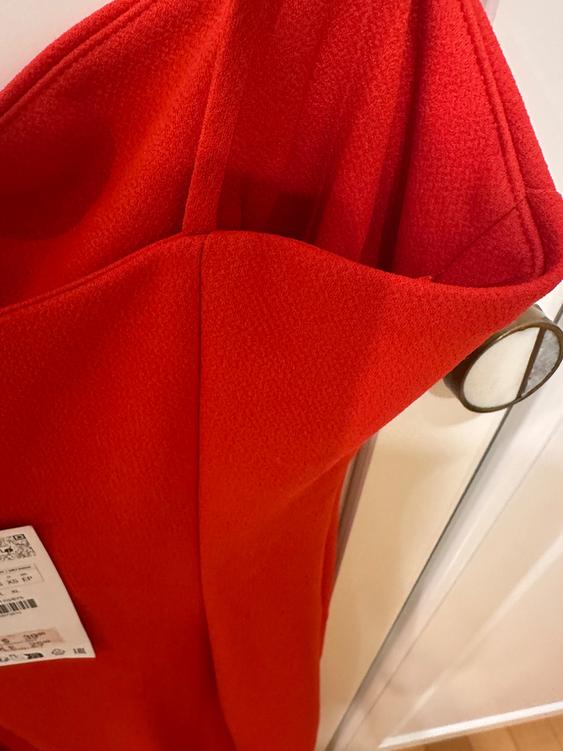 Red dress hanging on a white hanger, with visible tag. Textured fabric, warm and modern ambiance.