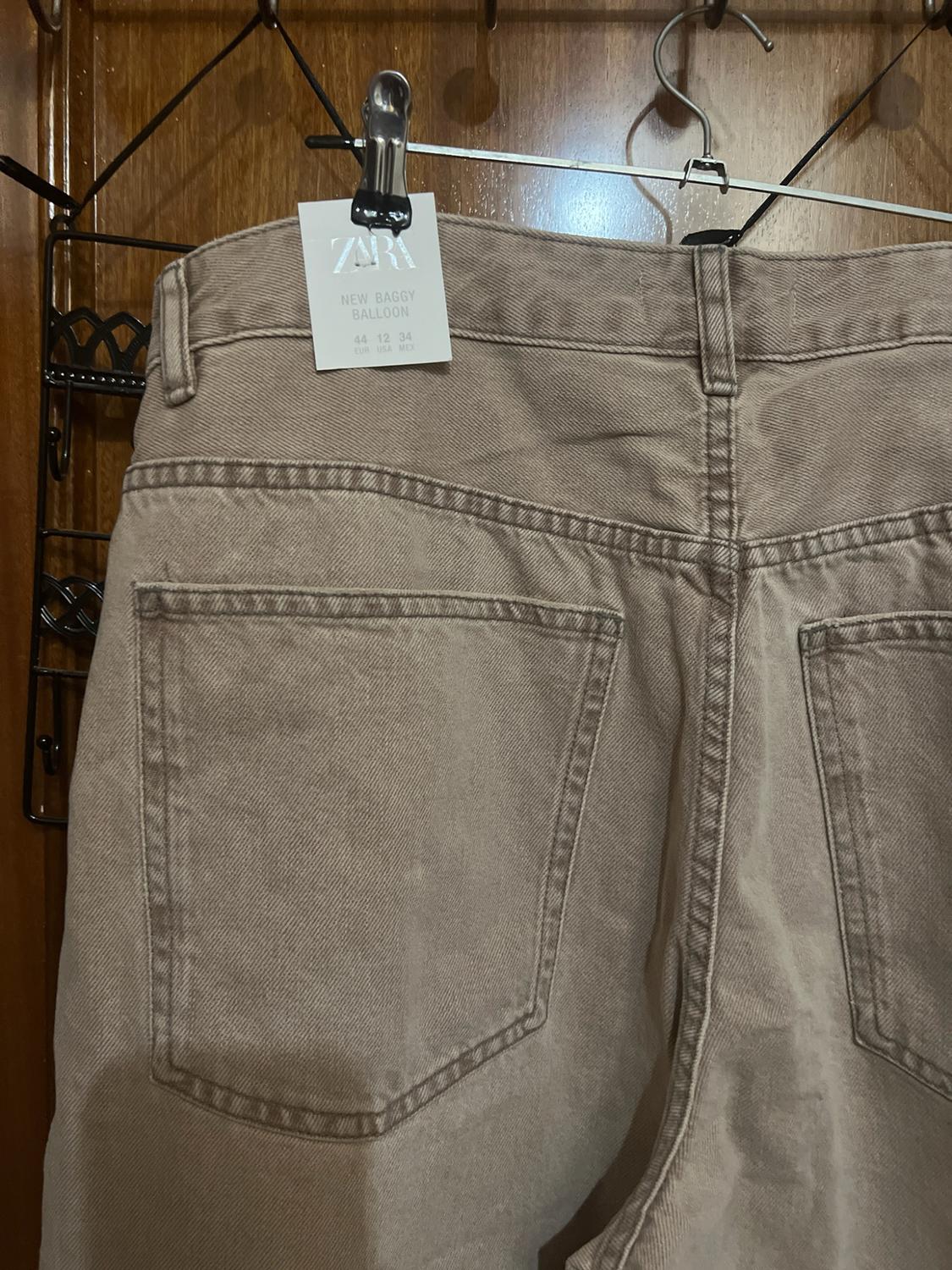Back of hanging beige jeans. Label: ZARA NEW BAGGY BALLOON 44 12 34 EUR USA MEX. Wooden background.