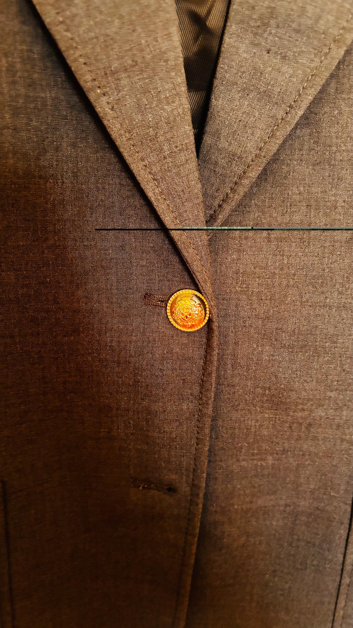 Detail of a brown blazer with a classic lapel and a decorative gold button in the centre. Visible textured fabric.