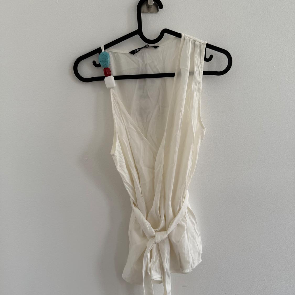 Sleeveless white blouse with a V-neck and adjustable belt, hanging on a black hanger against a white wall.