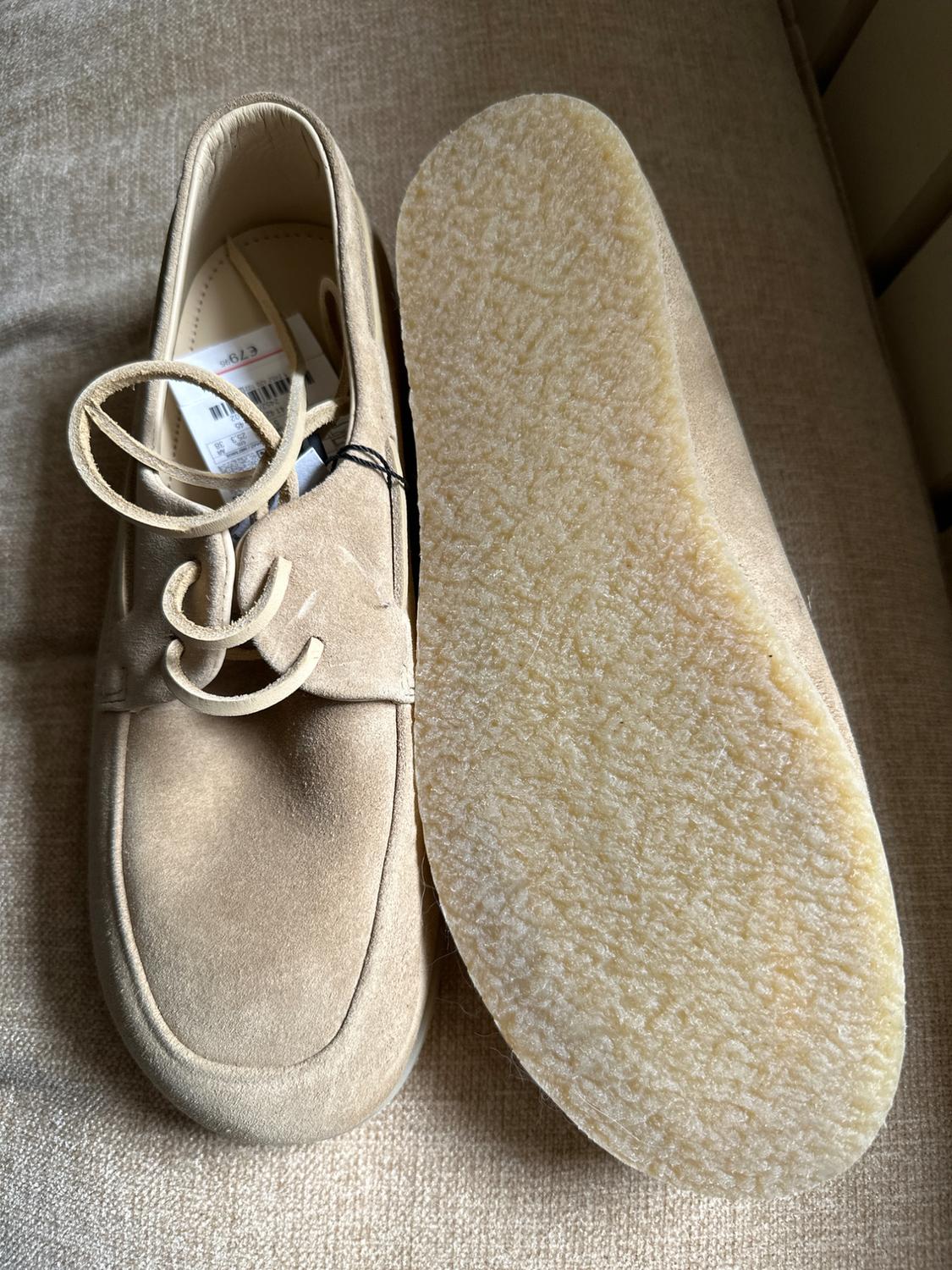 Pair of beige suede shoes on fabric. One shows laces and a tag with €79.95, the other is upside down showing the textured sole.