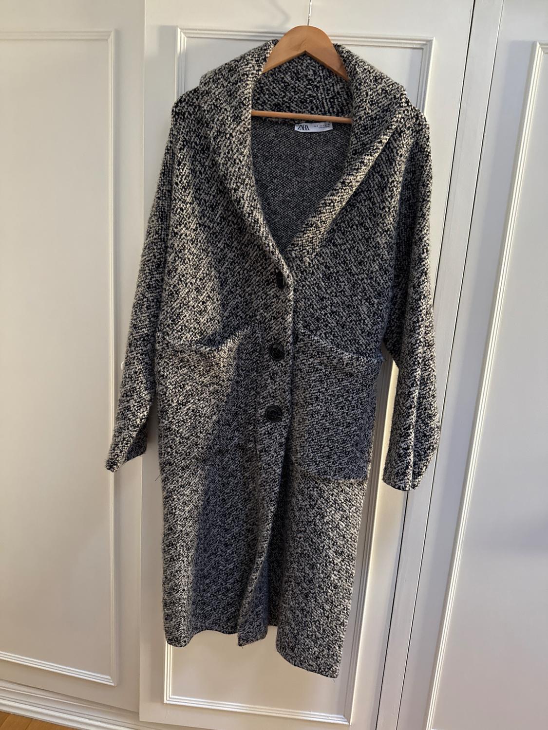 Chunky knit grey coat hanging on a wooden hanger, with large buttons and pockets, on a white door.