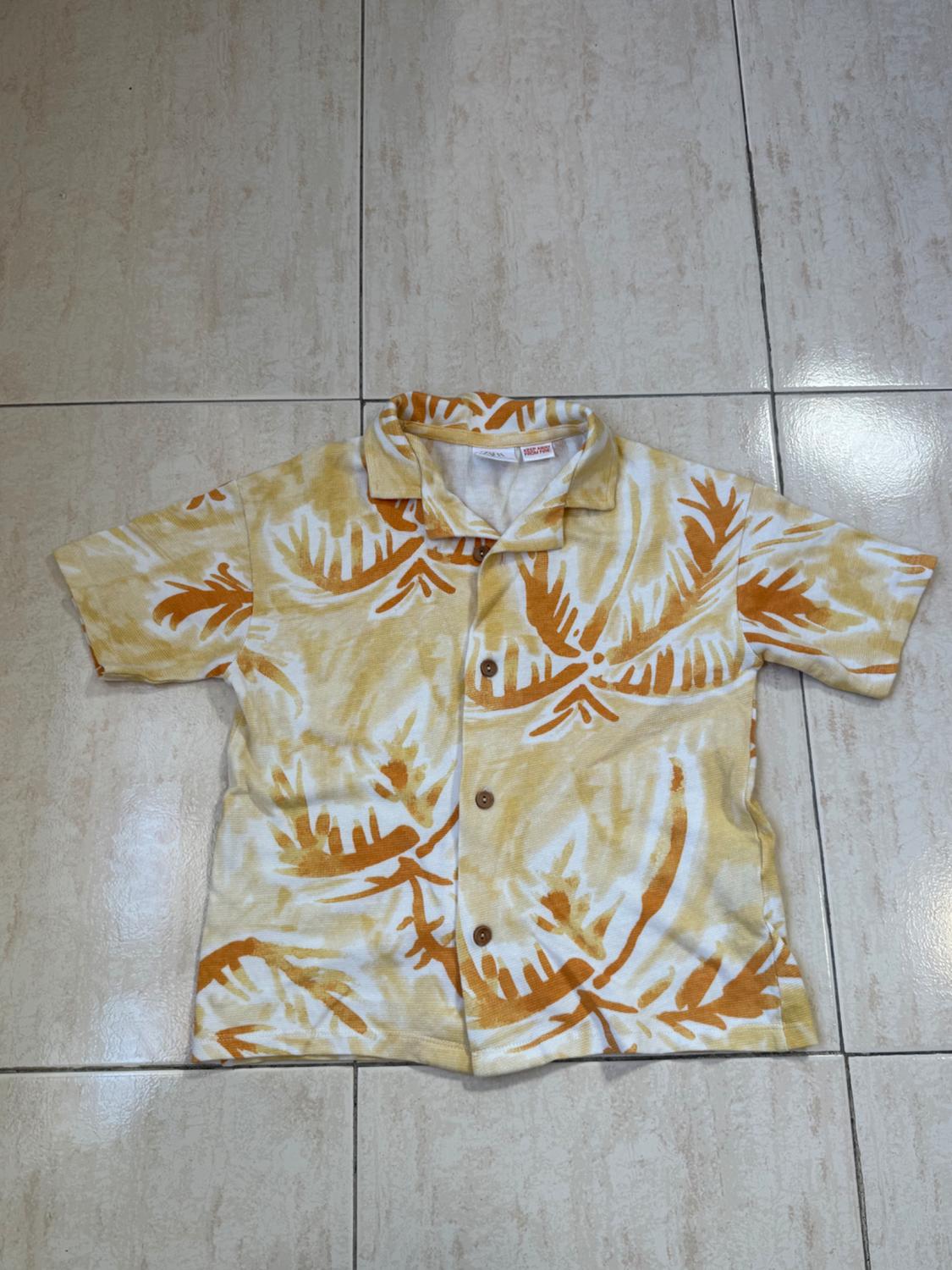 Short-sleeved shirt with a yellow and orange leaf print on a white background, placed on a light-coloured floor.