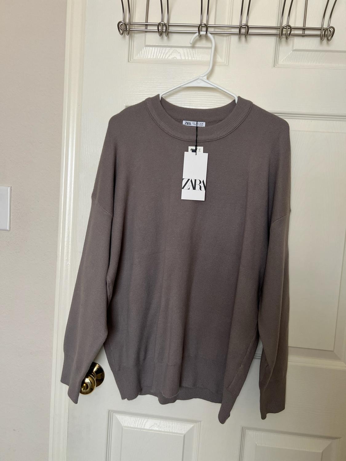 Brown knit sweater hanging on a hanger over a white door. Label with the text ZARA.