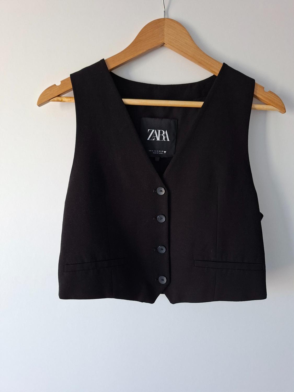 Black buttoned waistcoat on a wooden hanger against a white background. Label with the text ZARA EUR S USA S MEX 26 MADE IN TURKEY.